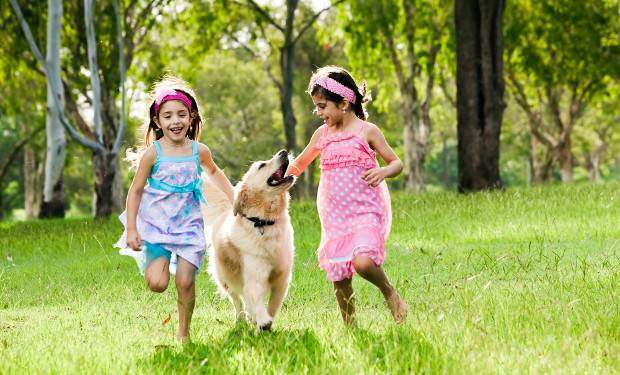 Two girls running with a golden retriever away from some trees across grass
